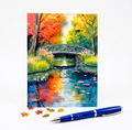 Colorful painting of a bridge over water with autumn trees on note card and a blue pen in front of card