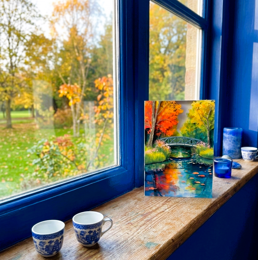 Autumn-themed note card of a bridge over water, on a windowsill with mugs and a blue candle.