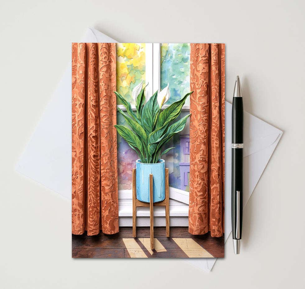 Note card with a peace lily plant in front of a window as subject. Card sitting on white surface with a pen next to card and envelope