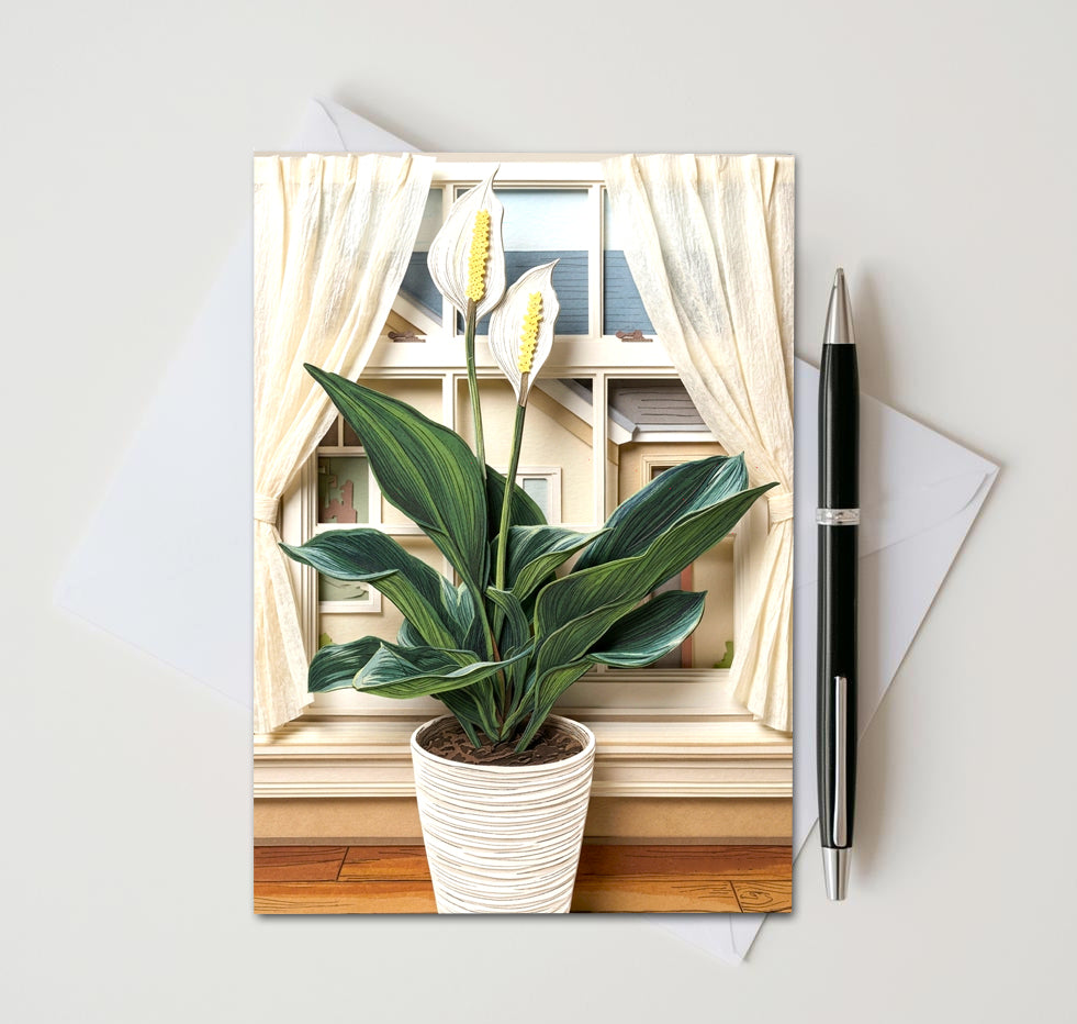 Note card with a peace lily plant in front of a window as subject. Card sitting on white surface with a pen next to card and envelope