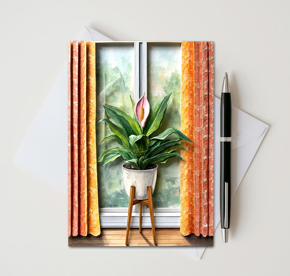 Note card with a peace lily plant in front of a window as subject. Card sitting on white surface with a pen next to card and envelope