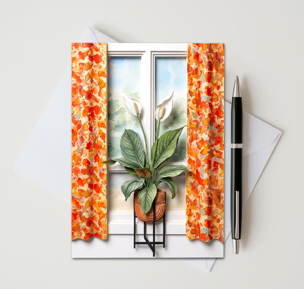Note card with a peace lily plant in front of a window as subject. Card sitting on white surface with a pen next to card and envelope