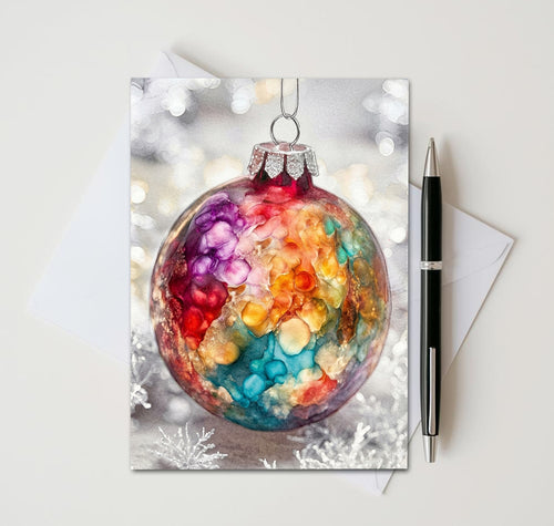 Christmas card with purple, red, gold, and teal bauble against snowy lights, beside an envelope and pen
