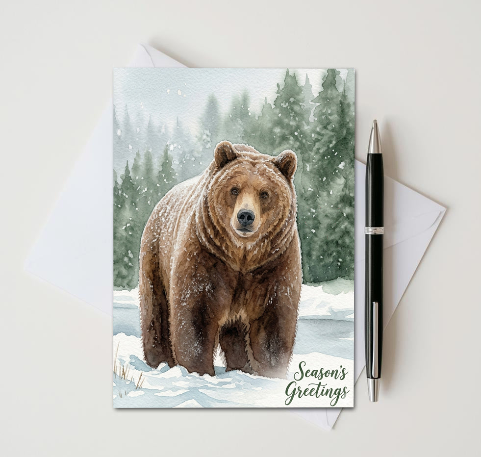 Season’s Greetings card with brown bear in a snowy evergreen forest, beside an envelope and pen

