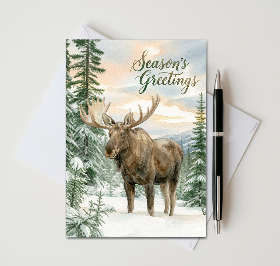 Season’s Greetings card with moose among snowy evergreen trees, beside an envelope and pen
